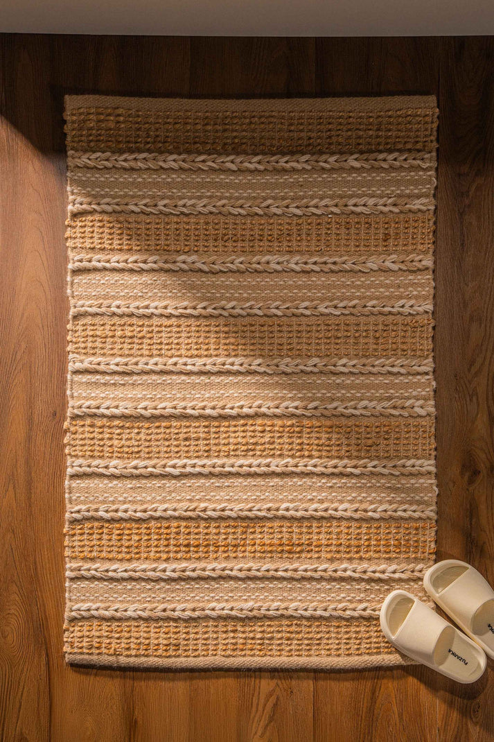 Tapete retangular de fibras naturais em tons de bege e marrom claro, com textura em listras horizontais e chinelos ao lado.