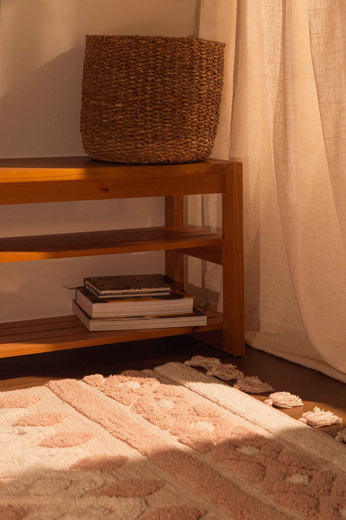 Cesto de palha em prateleira de madeira clara, com cortina bege ao lado. Tapete felpudo rosado com textura e franjas no chão.